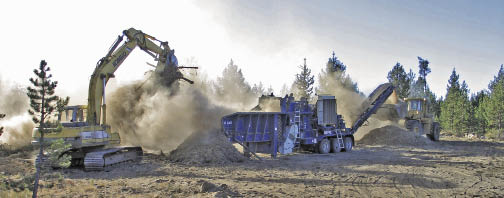 Figure 10.19 - Large mulching operations require access and working space. The operation shown in this photograph shows the wood waste material being dropped into a grinder with an excavator (left), and conveyed as mulch to the bucket of a front end loader (right).